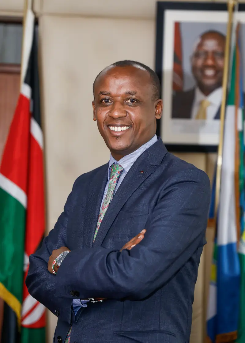 Mutula Kilonzo Jr. in a navy suit, with folded arms, beside the Kenyan and Makueni County flags.